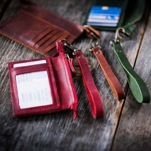 May include: A red leather wallet with a zipper closure and a clear plastic window for displaying an ID card. The wallet is attached to a red leather lanyard with a metal clasp. There are three other leather lanyards in green, brown, and orange.