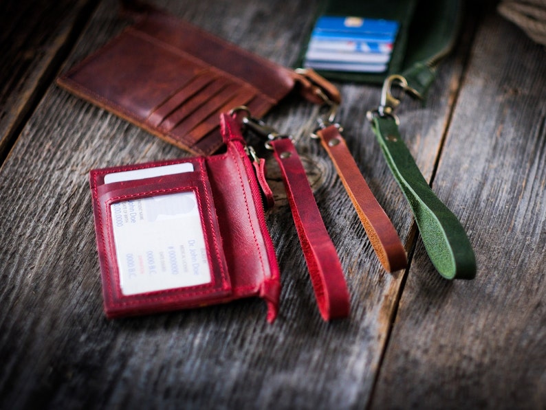 May include: A red leather wallet with a zipper closure and a clear plastic window for displaying an ID card. The wallet is attached to a red leather lanyard with a metal clasp. There are three other leather lanyards in green, brown, and orange.