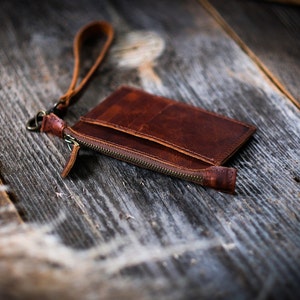 May include: A brown leather wallet with a zipper closure and a wrist strap. The wallet is lying on a wooden surface.