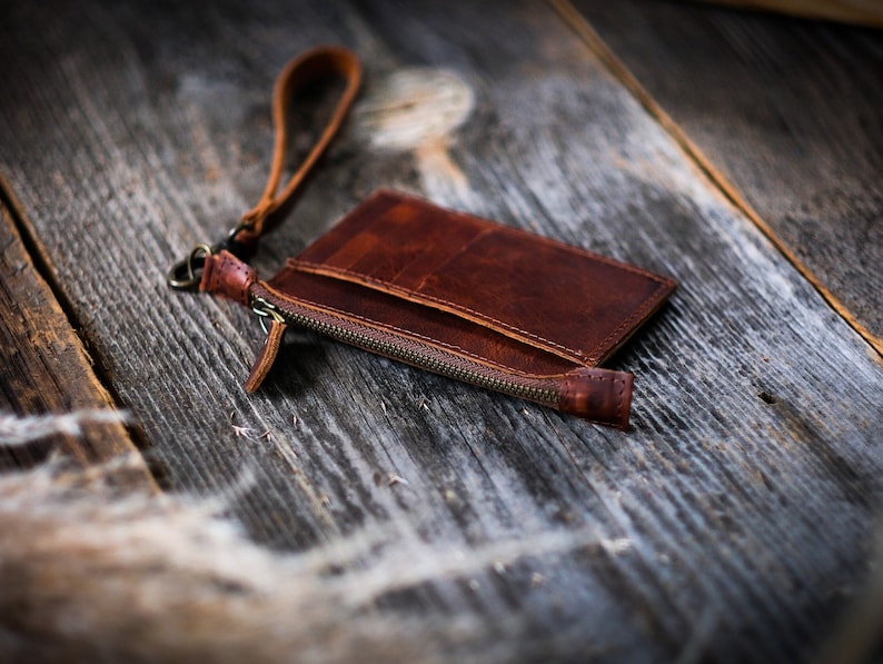 May include: A brown leather wallet with a zipper closure and a wrist strap. The wallet is lying on a wooden surface.