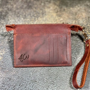 May include: A brown leather wallet with a zipper closure and a wrist strap. The wallet has a personalized inscription that reads "ACW".