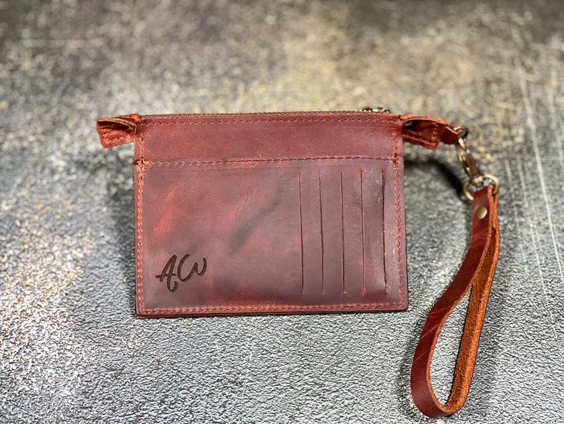 May include: A brown leather wallet with a zipper closure and a wrist strap. The wallet has a personalized inscription that reads "ACW".