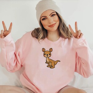 May include: A pink sweatshirt with a cartoon kangaroo graphic. The kangaroo is brown with a pink nose and cheeks.