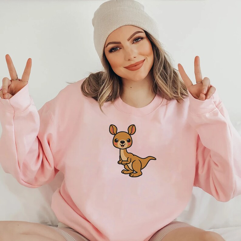 May include: A pink sweatshirt with a cartoon kangaroo graphic. The kangaroo is brown with a pink nose and cheeks.