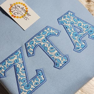May include: A light blue sweatshirt with the letters "ZTA" embroidered in a white and blue floral pattern.
