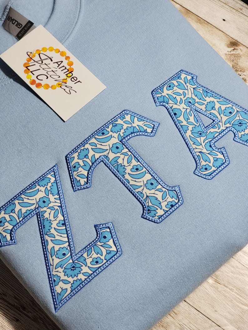 May include: A light blue sweatshirt with the letters "ZTA" embroidered in a white and blue floral pattern.