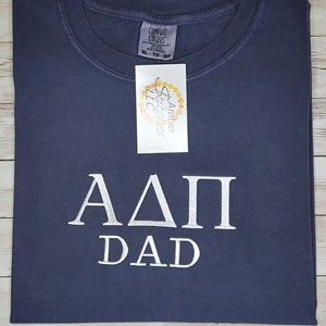 May include: A dark blue t-shirt with white lettering that says "ΑΔΠ DAD".