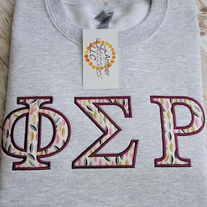 May include: A light gray sweatshirt with embroidered Greek letters ΦΣΡ in a burgundy outline. The letters are filled with a colorful leaf pattern. A tag from Amber Stitches LLC is attached.