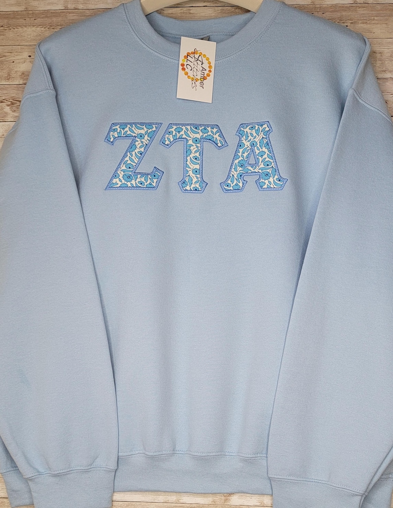 May include: Light blue sweatshirt with the letters "ZTA" embroidered in white with a blue floral pattern.