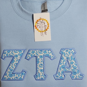 May include: Light blue sweatshirt with a white and blue floral pattern embroidered in the letters "ZTA". The sweatshirt has a tag that reads "Amber Stitches LLC".