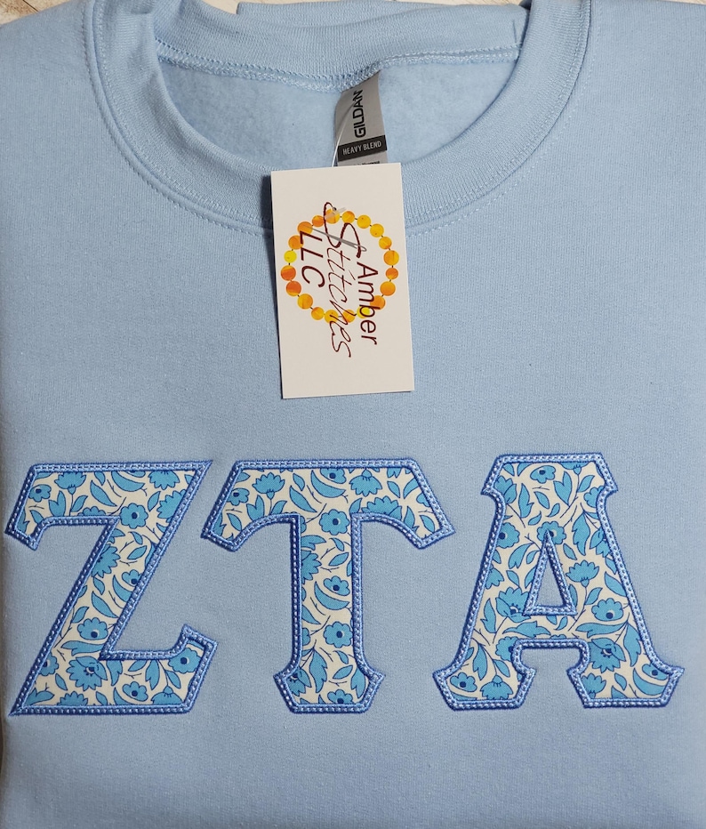 May include: Light blue sweatshirt with a white and blue floral pattern embroidered in the letters "ZTA". The sweatshirt has a tag that reads "Amber Stitches LLC".
