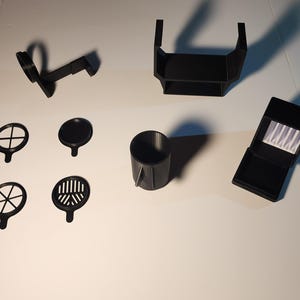 May include: A set of black 3D printed objects, including a small box with a white interior, a cylindrical container, a stand with a shelf, and four circular objects with different patterns.