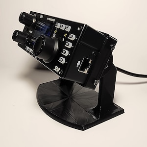 Puede incluir: Un transceptor SDR negro con múltiples perillas y botones, montado en un soporte negro. El dispositivo tiene una pequeña pantalla y el texto "SDR" es visible. También hay un micrófono y un puerto Ethernet. Un cable negro se extiende desde el dispositivo.