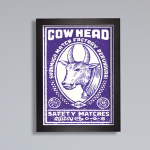 May include: A vintage matchbox label with a purple background and white text. The label reads "Cow Head Factory Perundurai Shumuga Match Safety Matches". A black and white illustration of a cow's head is in the center of the label.