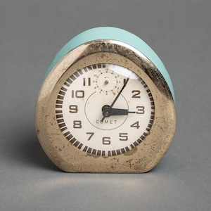May include: Vintage Comet alarm clock with a light blue top and a metallic face. The clock face is white with black numerals and hands, and the word "COMET" is printed below the center. The clock is set to approximately 3:15.