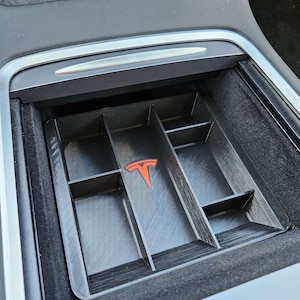 May include: A black plastic organizer with multiple compartments for a car's center console. The organizer has a red Tesla logo in the center.