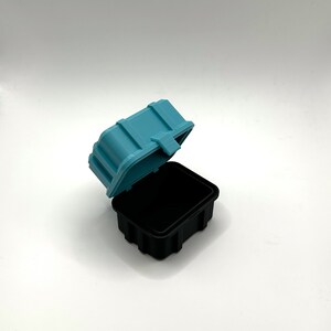 Pu&ograve; includere: Una piccola scatola di plastica blu turchese con un coperchio di plastica nero. Il coperchio &egrave; aperto e la scatola &egrave; vuota.