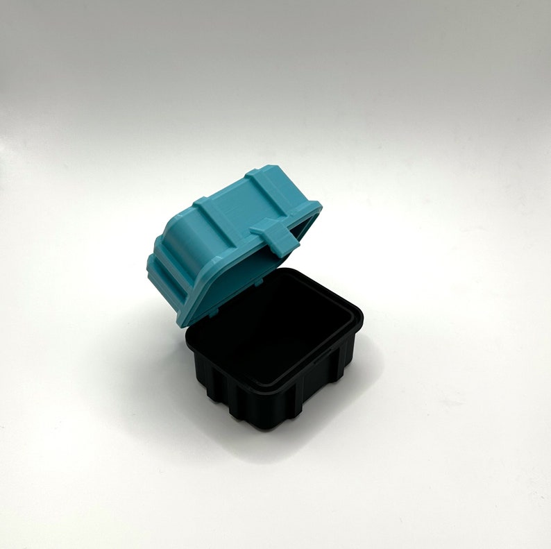Pu&ograve; includere: Una piccola scatola di plastica blu turchese con un coperchio di plastica nero. Il coperchio &egrave; aperto e la scatola &egrave; vuota.