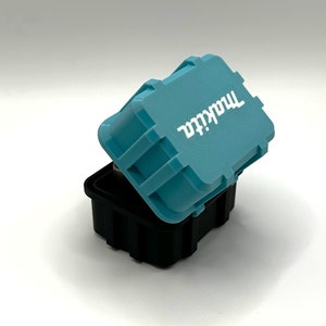 May include: A small, teal blue, plastic storage container with the word "Makita" printed in white on the lid. The container is open and the lid is slightly ajar.