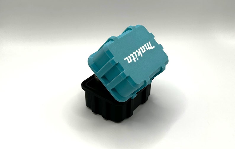 May include: A small, teal blue, plastic storage container with the word "Makita" printed in white on the lid. The container is open and the lid is slightly ajar.