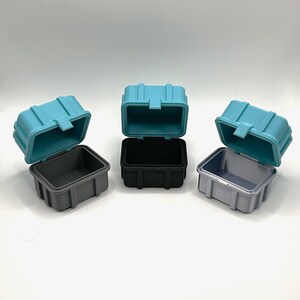 Pu&ograve; includere: Tre piccole scatole di immagazzinaggio in plastica aperte. Le scatole sono di colore turchese, nero e grigio.