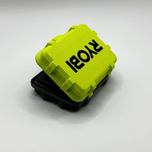 Pode incluir: Uma pequena caixa de armazenamento retangular de cor verde brilhante com uma tampa preta. A caixa tem a palavra "RYOBI" impressa em letras pretas na parte superior.