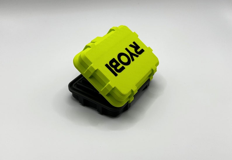 Pode incluir: Uma pequena caixa de armazenamento retangular de cor verde brilhante com uma tampa preta. A caixa tem a palavra "RYOBI" impressa em letras pretas na parte superior.