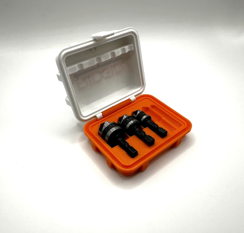 May include: A set of three drill bits in a white and orange plastic case. The drill bits are black and silver with a pointed tip.