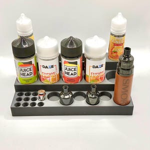 May include: A grey plastic organiser with six slots for bottles and 24 smaller slots for vape coils. The organiser holds six bottles of e-liquid, including Juice Head Strawberry Kiwi, Juice Head Pineapple Grapefruit, DAZE Fusion Honeydew, DAZE Fusion Peach Mango, and FINEST Grape Passion. A brown vape mod with the word "SMOK" printed on it is also in the organiser.