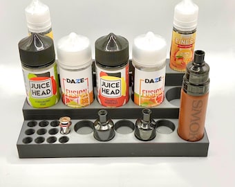 Op de afbeelding: Een grijze plastic organizer met zes vakken voor flessen en 24 kleinere vakken voor vape-coils. De organizer bevat zes flessen e-liquid, waaronder Juice Head Strawberry Kiwi, Juice Head Pineapple Grapefruit, DAZE Fusion Honeydew, DAZE Fusion Peach Mango en FINEST Grape Passion. Een bruine vape-mod met het woord "SMOK" erop is ook in de organizer.