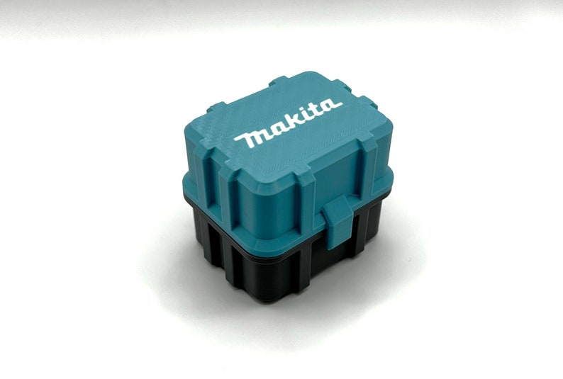 May include: A small teal blue plastic storage case with a black base. The case has a latch on the front and the word "Makita" printed in white on the top.