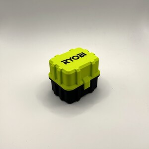K&ouml;nnte beinhalten: Eine kleine, leuchtend gr&uuml;ne Aufbewahrungsbox aus Kunststoff mit schwarzem Boden. Die Box hat einen Verschluss an der Vorderseite und das Wort "RYOBI" in Schwarz auf der Oberseite.