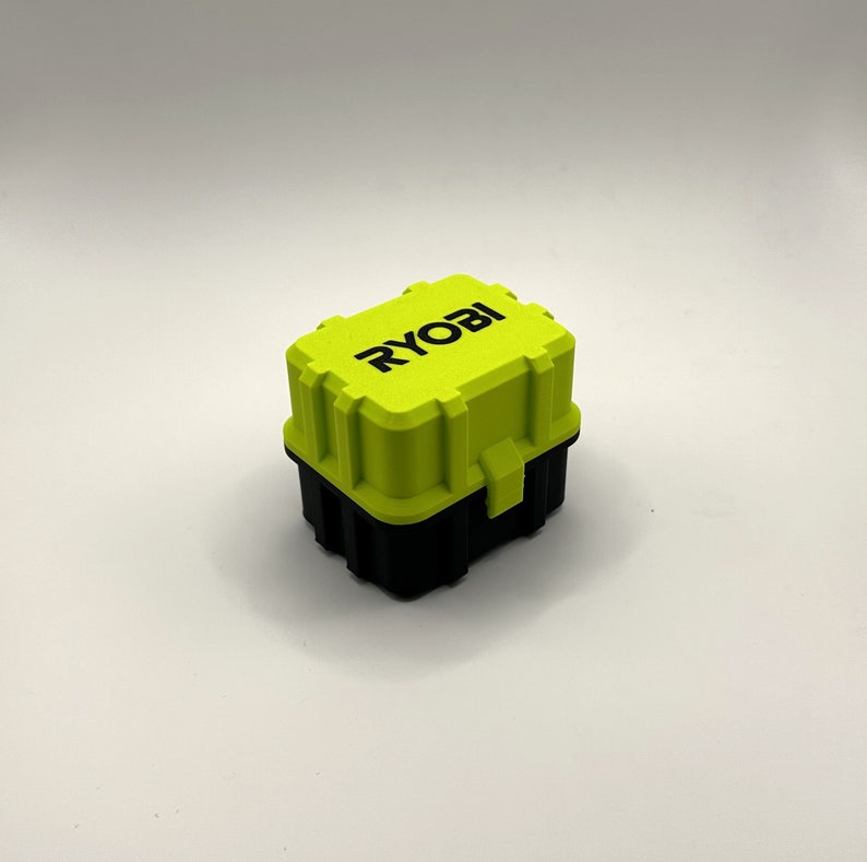K&ouml;nnte beinhalten: Eine kleine, leuchtend gr&uuml;ne Aufbewahrungsbox aus Kunststoff mit schwarzem Boden. Die Box hat einen Verschluss an der Vorderseite und das Wort "RYOBI" in Schwarz auf der Oberseite.