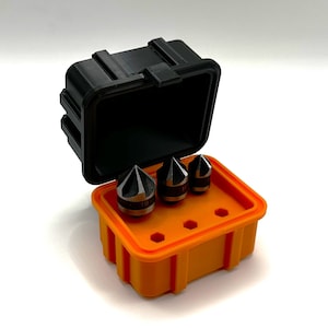 Op de afbeelding: Een set van drie zwarte en zilveren boortjes in een feloranje plastic koffer met een zwart deksel. De koffer heeft een label met het nummer "19" erop.