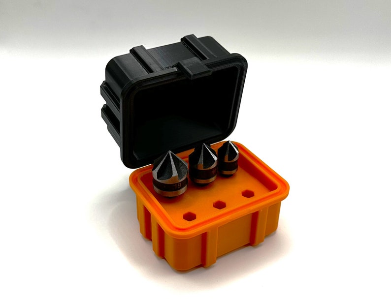 Op de afbeelding: Een set van drie zwarte en zilveren boortjes in een feloranje plastic koffer met een zwart deksel. De koffer heeft een label met het nummer "19" erop.
