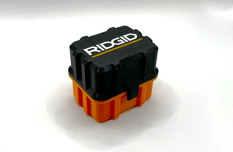 Op de afbeelding: Een kleine, zwarte en oranje plastic opbergdoos met het woord "RIDGID" in wit op de bovenkant.
