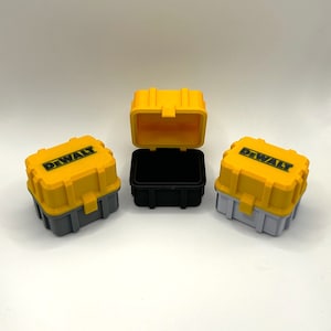 Könnte beinhalten: Drei kleine gelbe Werkzeugkästen mit dem DEWALT-Logo. Die Werkzeugkästen sind geöffnet und haben unterschiedlich farbige Innenräume: Schwarz, Grau und Weiß.