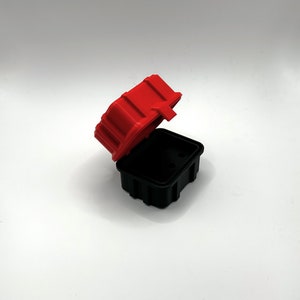 Op de afbeelding: Een klein rood en zwart plastic doosje met een scharnierend deksel. Het doosje is open en het deksel is opengeklapt.