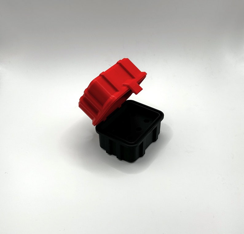 Op de afbeelding: Een klein rood en zwart plastic doosje met een scharnierend deksel. Het doosje is open en het deksel is opengeklapt.