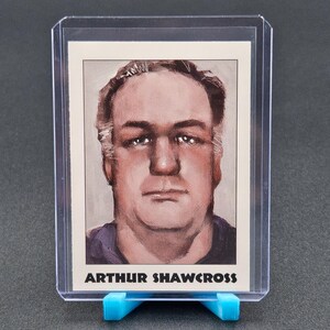 May include: A trading card featuring a painted portrait of a man with the name "ARTHUR SHAWCROSS" printed in black at the bottom. The card is encased in a clear plastic holder and displayed on a blue stand.