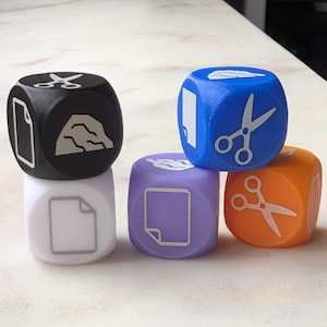 Peut inclure: Un ensemble de cinq dés multicolores arborant des icônes blanches de ciseaux, de papier et de pierre. Les dés sont noirs, blancs, bleus, violets et orange. Chaque face de dé affiche un symbole graphique clair représentant le jeu pierre-feuille-ciseaux.