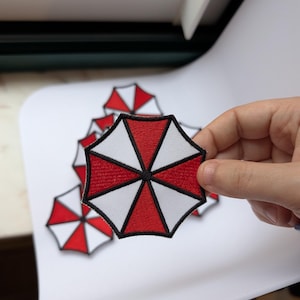 May include: A circular embroidered patch featuring a red and white umbrella design divided into eight alternating segments. The segments are separated by thin black lines, and the entire patch is outlined with a thick black border.