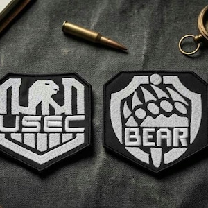 May include: Two black and white embroidered patches with shield designs. One patch reads "USEC" with a bear silhouette, the other reads "BEAR" with a bear claw design. A bullet and a compass are also visible.