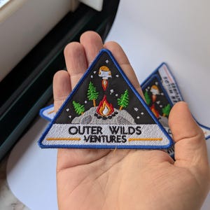 May include: Embroidered triangular patch with a black background, featuring a rocket ship, campfire, trees, and the text "OUTER WILDS VENTURES". The patch has a blue border and is held in a hand.