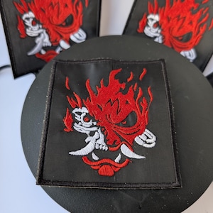 Puede incluir: Parche cuadrado negro con un diseño de calavera en llamas rojas y detalles blancos. El parche, probablemente bordado, está sobre un fondo negro. Adecuado para ropa o accesorios.