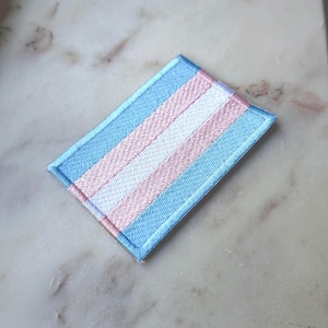Op de afbeelding: Een rechthoekige geborduurde patch met een streeppatroon in lichtblauw, lichtroze en wit. Het ontwerp bestaat uit twee lichtblauwe buitenste banen, twee lichtroze binnenste banen en een enkele witte centrale baan met gestikte randen.