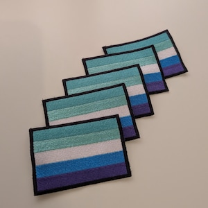 Puede incluir: Cinco parches rectangulares bordados dispuestos en una fila diagonal. Cada parche presenta cinco franjas horizontales en tonos verde azulado, verde claro, blanco, azul medio y morado oscuro, encerrados en un borde negro cosido.