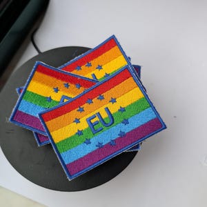 Peut inclure: Écussons brodés avec un drapeau arc-en-ciel, des étoiles bleues et les lettres "EU". Les écussons sont rectangulaires avec une bordure bleue. Les couleurs de l'arc-en-ciel comprennent le rouge, l'orange, le jaune, le vert, le bleu et le violet.