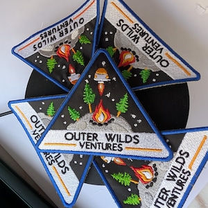 May include: Triangular embroidered patches with the text "Outer Wilds Ventures." The patches feature a black background with a rocket ship, flames, trees, and a blue border. The patches are white, orange, green, and blue.