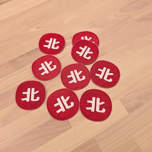May include: Eight circular red fabric patches, each featuring the white Japanese kanji character for north, 北, embroidered in the centre. The patches are arranged in a cluster, showcasing a consistent round shape and a stitched border design.